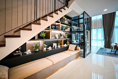A modern interior space features an under-staircase shelving unit adorned with decorative items and indoor plants. The space includes a beige cushioned bench, a glass-encased lift, and a large window with sheer curtains allowing natural light. A cozy atmosphere is complemented by contemporary furniture and decor.