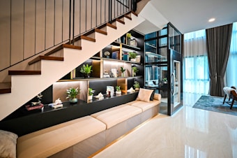 A modern interior space features an under-staircase shelving unit adorned with decorative items and indoor plants. The space includes a beige cushioned bench, a glass-encased lift, and a large window with sheer curtains allowing natural light. A cozy atmosphere is complemented by contemporary furniture and decor.
