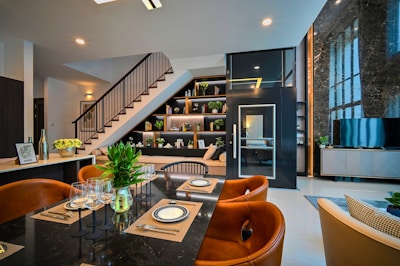 A modern and stylish interior featuring a dining area with a black marble table set with plates, cutlery, and wine glasses. Brown leather chairs surround the table, and there's a vase with green plants as a centerpiece. In the background, an elegant staircase with dark railing leads to a loft. Shelves beneath the stairs display books and decorative items. A television is positioned on a sleek console near a large marble-featured wall with expansive windows.