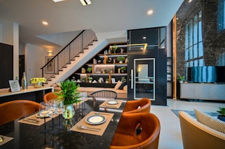 A modern and stylish interior featuring a dining area with a black marble table set with plates, cutlery, and wine glasses. Brown leather chairs surround the table, and there's a vase with green plants as a centerpiece. In the background, an elegant staircase with dark railing leads to a loft. Shelves beneath the stairs display books and decorative items. A television is positioned on a sleek console near a large marble-featured wall with expansive windows.