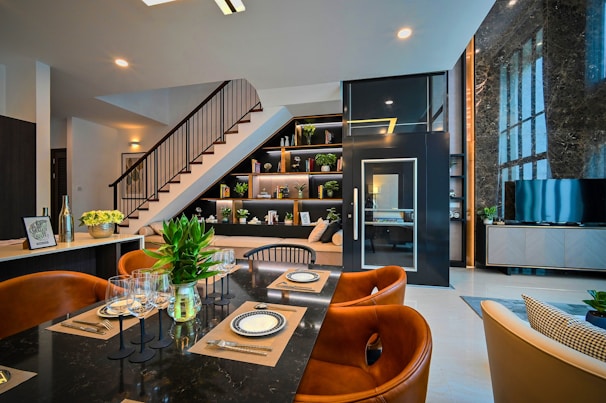 A modern and stylish interior featuring a dining area with a black marble table set with plates, cutlery, and wine glasses. Brown leather chairs surround the table, and there's a vase with green plants as a centerpiece. In the background, an elegant staircase with dark railing leads to a loft. Shelves beneath the stairs display books and decorative items. A television is positioned on a sleek console near a large marble-featured wall with expansive windows.