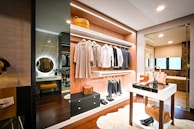 A modern walk-in closet with an open display of clothing items hanging neatly on racks. The closet features wooden shelves, drawers, and a mirror. On the floor, there is a soft, white rug. A small table holds fashion accessories such as bags and shoes. The lighting creates a cozy and organized atmosphere.