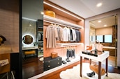 A modern walk-in closet with an open display of clothing items hanging neatly on racks. The closet features wooden shelves, drawers, and a mirror. On the floor, there is a soft, white rug. A small table holds fashion accessories such as bags and shoes. The lighting creates a cozy and organized atmosphere.