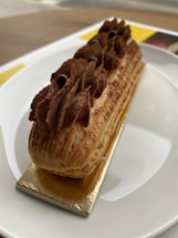 A delicate pastry topped with rich chocolate cream is set on a rectangular golden base, resting elegantly on a white plate. The pastry has a textured, light brown exterior with piped chocolate cream adorned with small, dark chocolate beads.
