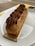 A delicate pastry topped with rich chocolate cream is set on a rectangular golden base, resting elegantly on a white plate. The pastry has a textured, light brown exterior with piped chocolate cream adorned with small, dark chocolate beads.