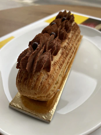 A delicate pastry topped with rich chocolate cream is set on a rectangular golden base, resting elegantly on a white plate. The pastry has a textured, light brown exterior with piped chocolate cream adorned with small, dark chocolate beads.