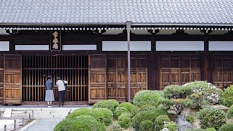Two people stand in front of a traditional wooden building with a tiled roof. The entrance is marked by wooden sliding doors and a horizontal sign with characters above it. Lush, manicured shrubs and a stone pathway lead up to the entrance, creating a serene garden setting.