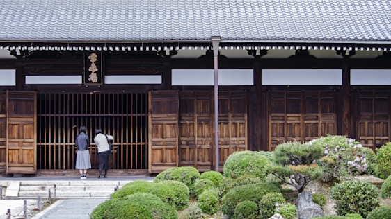 Two people stand in front of a traditional wooden building with a tiled roof. The entrance is marked by wooden sliding doors and a horizontal sign with characters above it. Lush, manicured shrubs and a stone pathway lead up to the entrance, creating a serene garden setting.