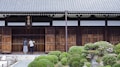 Two people stand in front of a traditional wooden building with a tiled roof. The entrance is marked by wooden sliding doors and a horizontal sign with characters above it. Lush, manicured shrubs and a stone pathway lead up to the entrance, creating a serene garden setting.