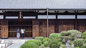 Two people stand in front of a traditional wooden building with a tiled roof. The entrance is marked by wooden sliding doors and a horizontal sign with characters above it. Lush, manicured shrubs and a stone pathway lead up to the entrance, creating a serene garden setting.