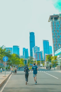 People are jogging on a city street with skyscrapers in the background. The atmosphere is lively, with several people walking, cycling, or engaging in various activities. Green trees line the street, adding a touch of nature to the urban environment.