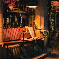 A cozy outdoor seating area featuring a wooden bench stool, a woven decorative basket, and a comfortable loveseat under soft string lights.