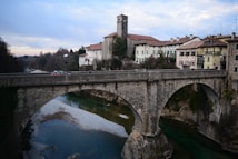 An old stone bridge spans a river surrounded by a serene landscape. The water below is a mix of turquoise and deep green, bordered by rocky shores. In the background, there are historic buildings with red-tiled roofs and a church tower, set against a backdrop of trees and a cloudy sky.