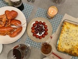 A dining table is set with an assortment of dishes including a large cooked lobster on a white plate, a bowl with a mix of lychees and cherries, and a baked cheesy lasagna in a glass dish. Additional elements on the table include wine glasses and a lit candle for ambiance.