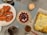 A dining table is set with an assortment of dishes including a large cooked lobster on a white plate, a bowl with a mix of lychees and cherries, and a baked cheesy lasagna in a glass dish. Additional elements on the table include wine glasses and a lit candle for ambiance.