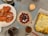 A dining table is set with an assortment of dishes including a large cooked lobster on a white plate, a bowl with a mix of lychees and cherries, and a baked cheesy lasagna in a glass dish. Additional elements on the table include wine glasses and a lit candle for ambiance.