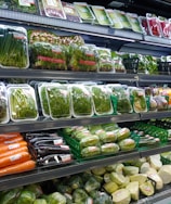 Various fresh vegetables packaged in plastic wrap and arranged neatly on supermarket shelves, including carrots, eggplants, cucumbers, and leafy greens.