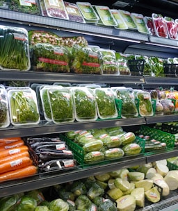 Various fresh vegetables packaged in plastic wrap and arranged neatly on supermarket shelves, including carrots, eggplants, cucumbers, and leafy greens.