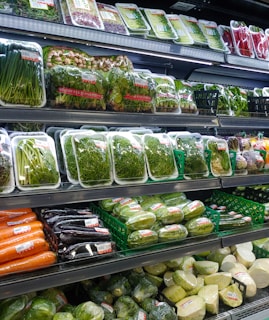 Various fresh vegetables packaged in plastic wrap and arranged neatly on supermarket shelves, including carrots, eggplants, cucumbers, and leafy greens.