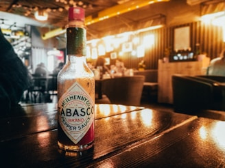 A Tabasco sauce bottle sits on a wooden table in a softly lit restaurant or cafe. The background features a warm, ambient glow with blurred seating areas and decor. The atmosphere appears cozy and inviting, with a focus on the bottle in the foreground.