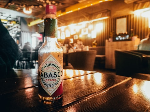 A Tabasco sauce bottle sits on a wooden table in a softly lit restaurant or cafe. The background features a warm, ambient glow with blurred seating areas and decor. The atmosphere appears cozy and inviting, with a focus on the bottle in the foreground.