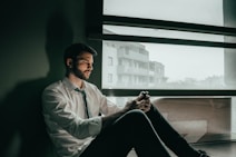 a man sitting on a window sill looking at his cell phone