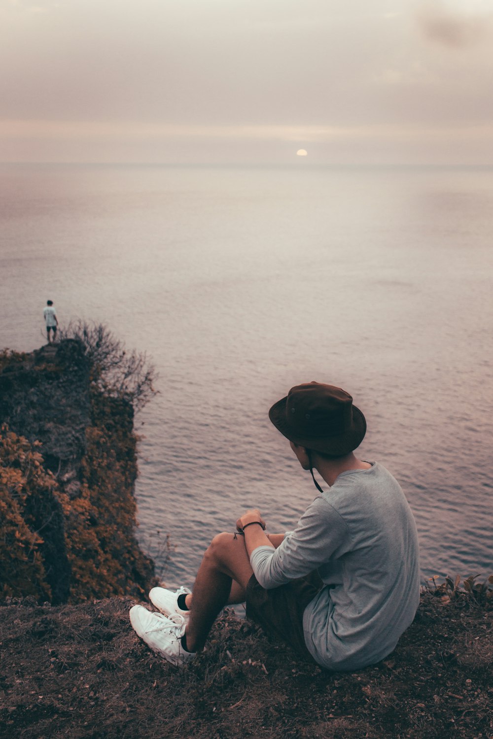 uomo in camicia grigia a maniche lunghe e cappello nero seduto sulla roccia vicino allo specchio d'acqua