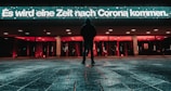 A person stands alone on a wet paved surface at night, facing a brightly lit building with red and white lights. A large illuminated sign in German reads 'Es wird eine Zeit nach Corona kommen.' The atmosphere appears calm yet reflective.