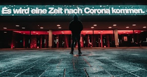 A person stands alone on a wet paved surface at night, facing a brightly lit building with red and white lights. A large illuminated sign in German reads 'Es wird eine Zeit nach Corona kommen.' The atmosphere appears calm yet reflective.