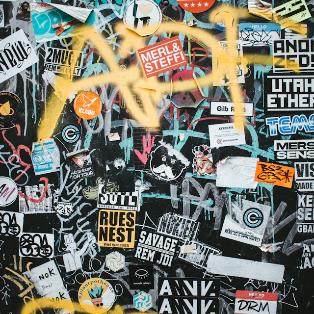 A dark wall covered in numerous colorful stickers and yellow spray paint graffiti
