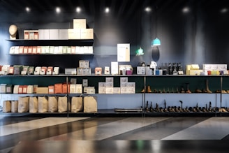 A well-organized display of various packaged goods and products on shelves. The top shelves hold neatly stacked boxes and packages of different colors, while the bottom shelves contain bags, shoes, and other items. Small pendant lights hang above with a soft green glow, and the overall setting has a modern and minimalist aesthetic.