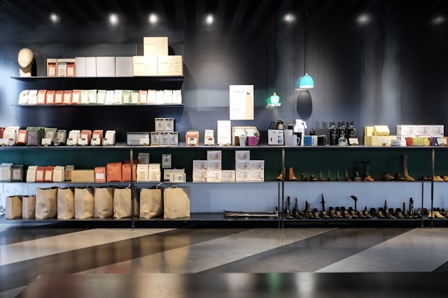 A well-organized display of various packaged goods and products on shelves. The top shelves hold neatly stacked boxes and packages of different colors, while the bottom shelves contain bags, shoes, and other items. Small pendant lights hang above with a soft green glow, and the overall setting has a modern and minimalist aesthetic.