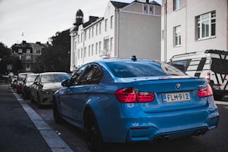 A blue BMW car is parked on the side of a street in an urban area. The car features a Finnish license plate and is positioned in front of several other vehicles, including a silver car and a van. The background consists of multi-story residential buildings with white and light-colored facades, as well as trees lining the street.