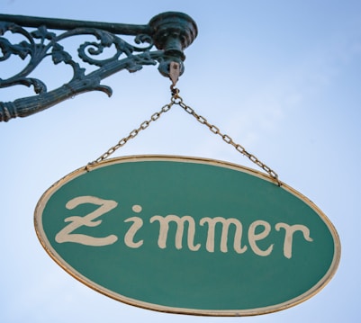 A vintage-style sign with ornate metalwork holding an oval-shaped plaque displaying the word 'Zimmer' in a classic font. The sign is against a clear blue sky.