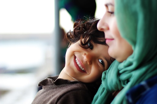 A young child with curly hair is smiling warmly while leaning against a woman wearing a green headscarf. They both share a close, affectionate moment, with the woman's face in profile.