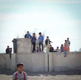 Several children are gathered on and around a large concrete structure outdoors. Some are standing while others are seated, appearing to be engaged in conversation or observing their surroundings. Their expressions vary, ranging from curious to relaxed.
