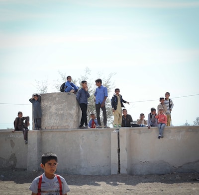 Several children are gathered on and around a large concrete structure outdoors. Some are standing while others are seated, appearing to be engaged in conversation or observing their surroundings. Their expressions vary, ranging from curious to relaxed.