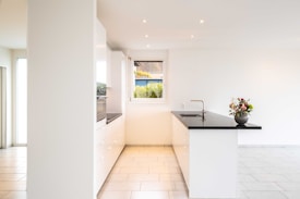 A modern, minimalist kitchen with white cabinetry and a black countertop. The kitchen island features a built-in sink and a vase with flowers. Soft ambient lighting illuminates the space. A window provides natural light and shows a glimpse of greenery outside.