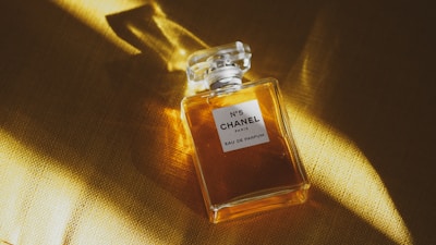 A clear glass bottle of Chanel No. 5 perfume with a square stopper rests on a golden surface. Light subtly illuminates the bottle, casting soft shadows and highlighting the amber liquid inside.