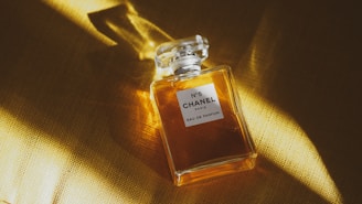 A clear glass bottle of Chanel No. 5 perfume with a square stopper rests on a golden surface. Light subtly illuminates the bottle, casting soft shadows and highlighting the amber liquid inside.