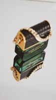 A rectangular green perfume bottle with a reflective surface and intricate gold chain decoration. The bottle is elegantly designed, featuring a textured black and gold pattern on the cap.