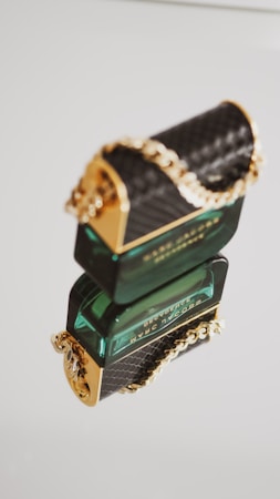 A rectangular green perfume bottle with a reflective surface and intricate gold chain decoration. The bottle is elegantly designed, featuring a textured black and gold pattern on the cap.