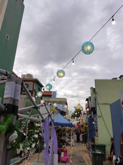 A vibrant street scene during a festive event, featuring strings of colorful lanterns hanging overhead against a cloudy sky. The street is lined with various stalls and decorations, including floral arrangements and ribbon streamers. There are people milling around, contributing to a lively atmosphere.