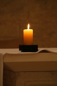 A single lit candle sits on a dark holder atop a flat, light-colored surface. The background is softly blurred, emphasizing the warm glow of the candle flame.