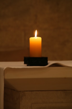A single lit candle sits on a dark holder atop a flat, light-colored surface. The background is softly blurred, emphasizing the warm glow of the candle flame.
