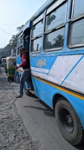 A person is seen stepping onto a blue and white public bus on a street. The bus has large windows and a design with text on its side. In the background, an auto-rickshaw and some trees are visible.