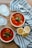 Two bowls of vegetable soup garnished with fresh herbs are on a wooden table next to sliced lemons and metal spoons. A striped cloth is spread out beneath the bowls.