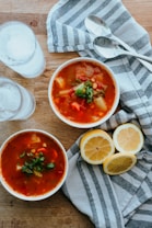 Two bowls of vegetable soup garnished with fresh herbs are on a wooden table next to sliced lemons and metal spoons. A striped cloth is spread out beneath the bowls.