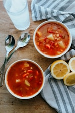 Two bowls of vegetable soup displayed on a wooden table, surrounded by slices of lemon and a striped cloth. Beside the bowls, two spoons and a glass of water can be seen, adding to the setting's simplicity and homeliness.