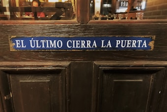 A wooden door with an aged blue sign that reads 'EL &Uacute;LTIMO CIERRA LA PUERTA' in white letters, situated in a dimly lit environment. A glimpse of an indoor setting with tables and glasses is visible through a window above the door.